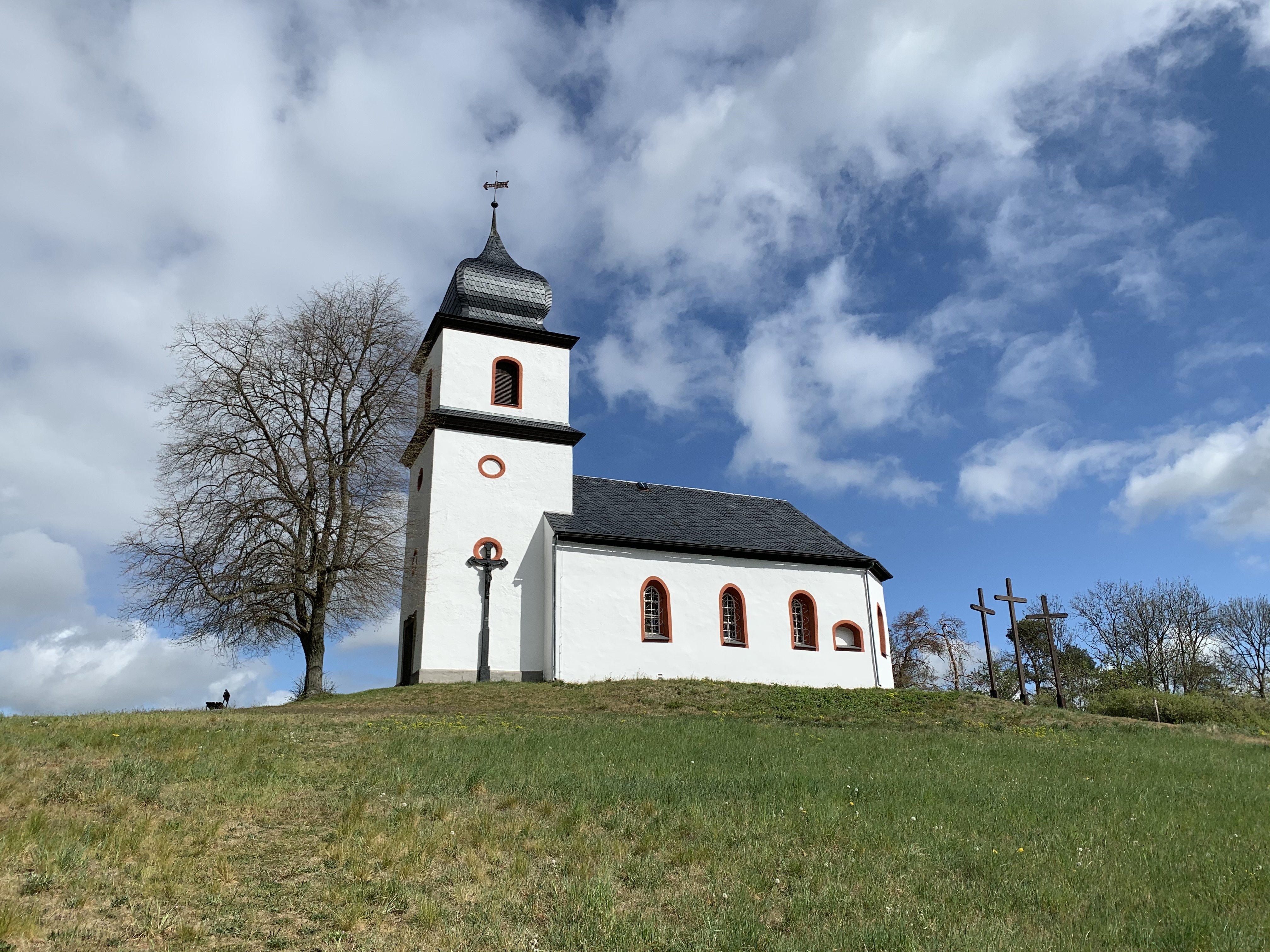 Kapelle auf grasbewachsenem Hgel unter Himmel. Kirchenhaus zwischen einzelnen Bumen und 3 Kreuzen.