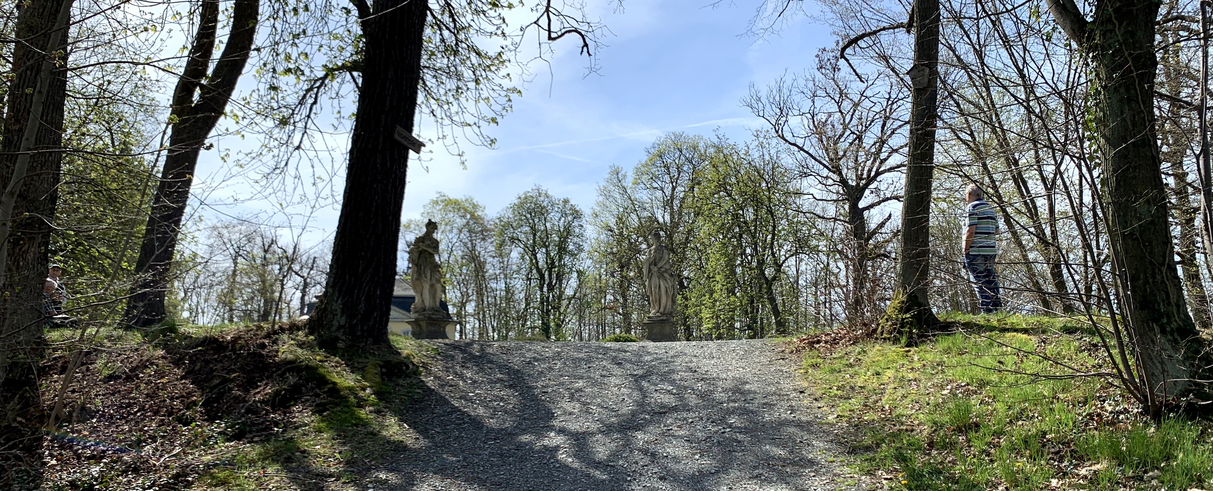 Wanderweg nach oben im Wald, endend bei einer Lichtung. Auf dem Weg stehen Statuen. Ein Mann steht und staunt bedächtig.