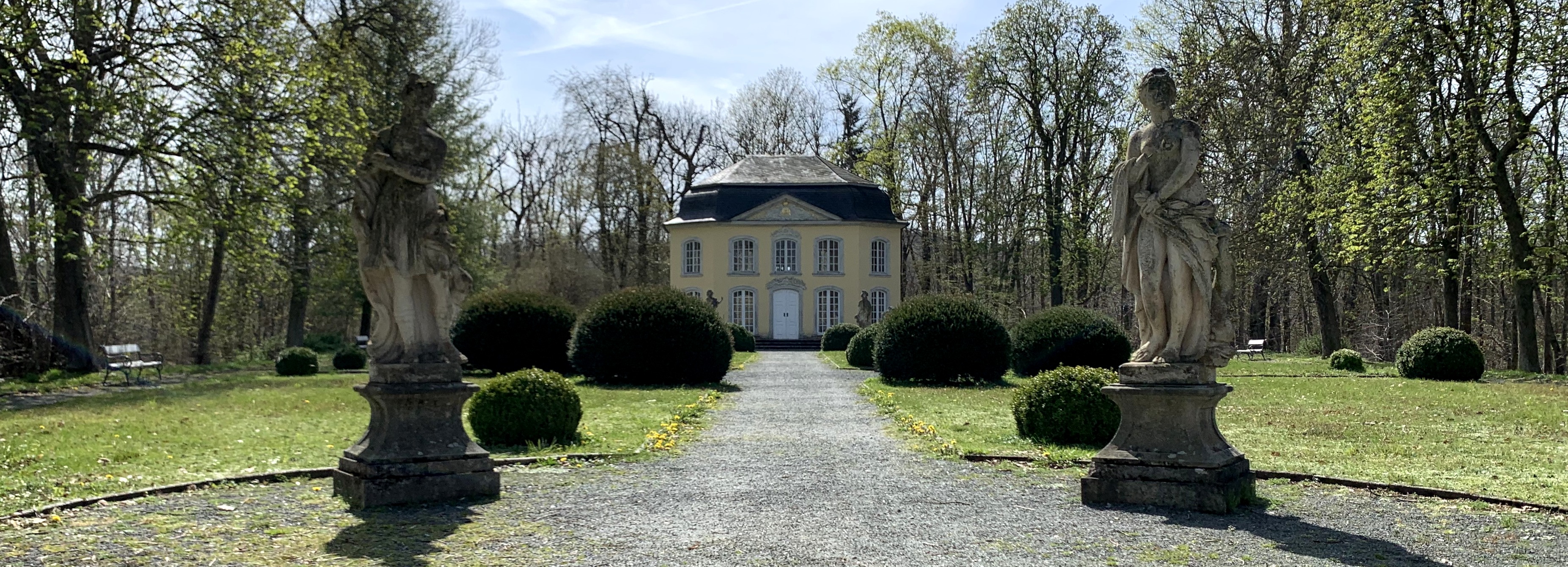 Schlosspark, Weg durch gemähte Wiese, gepflegte Büsche, zwischen Bäumen. Weg führt zu gelbem Häuschen. Weg beginbnt mit zwei Statuen, je eine auf jeder Seite