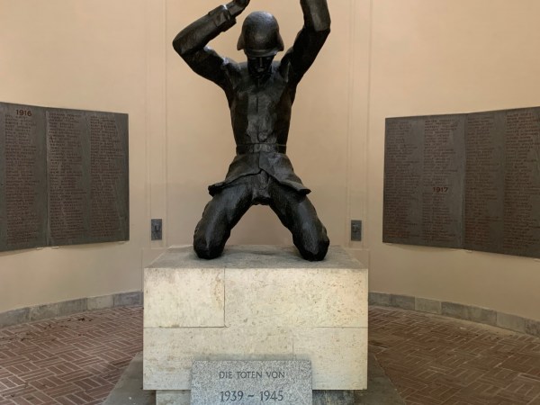 Auf einem Steinpodest Statue eines gefallenen Soldaten. An den Wänden Tafeln mit Namen. Steintafel und Blumen.