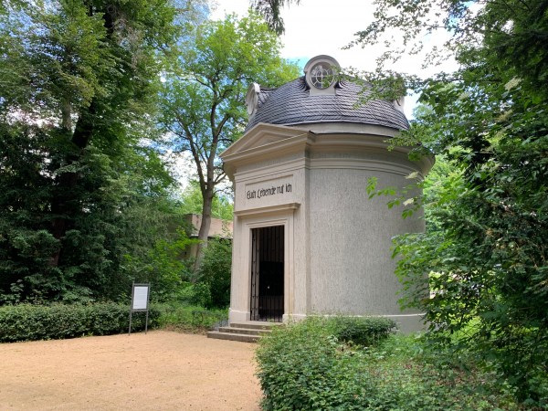 Runde Steinhütte im Park mit Gittertür. - Aufschrift: Euch Lebende ruf ich. Im Schieferdach zwei runde Fenster.