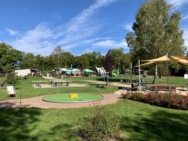 Bei schönem Wetter: Wiese mit Sträuchern, Sonnenschirmen, Minigolf, Tischtennisplatten und Bäumen unter leicht bewölktem blauem Himmel