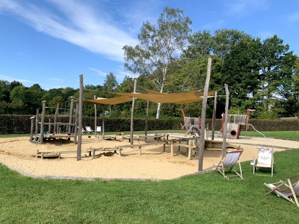 Sandkasten in Wiese vor Hecke vor Bäumen bei sonnigem Wetter. Sonnensegel überspannen den Sandkasten. Liegestühle stehen daneben.