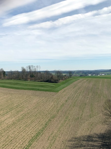 Von einer Aussichtsplattform Aussicht auf Feld. In der Ferne Wiesen, Bäume und Siedlungen. Alles unter leicht bewölktem Himmel.