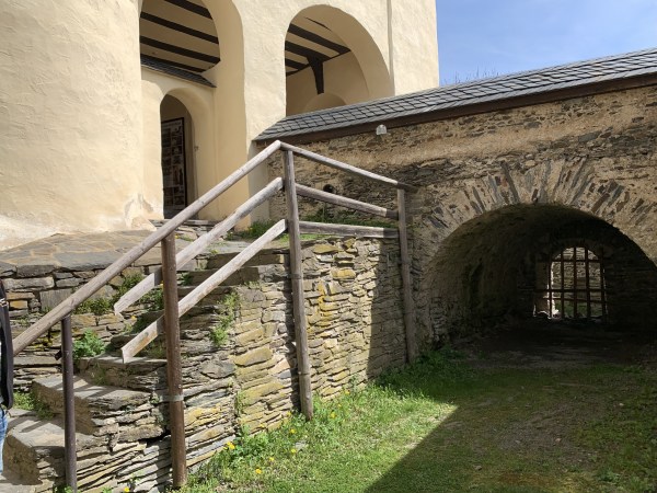 Steinerne Treppe mit Holzgeländer neben steinernem Torbogen. Am Ende des Torbogens im Dunklen ein Gitter. Steinmauer vom Torbogen mit schwarzem Schieferdach. Gebäude links mit beiger Wandfarbe.