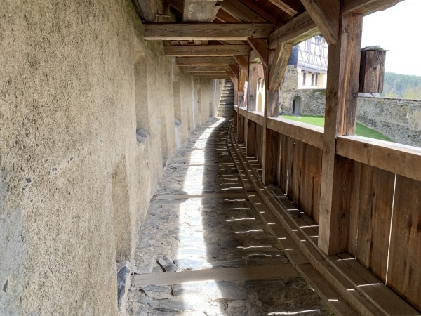 Verteidigungsgang zwischen Mauer mit Öffnungen und Innenhof. vor dem Innenhof Holzgeländer und Holzstützbalken.