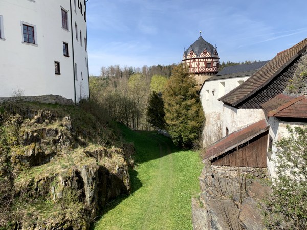 Schmaler Pfad zwischen weißen Haus auf Felsen und weißer Burgmauer. Vorbei an Burgturm. Bewachsen mit Bäumen.