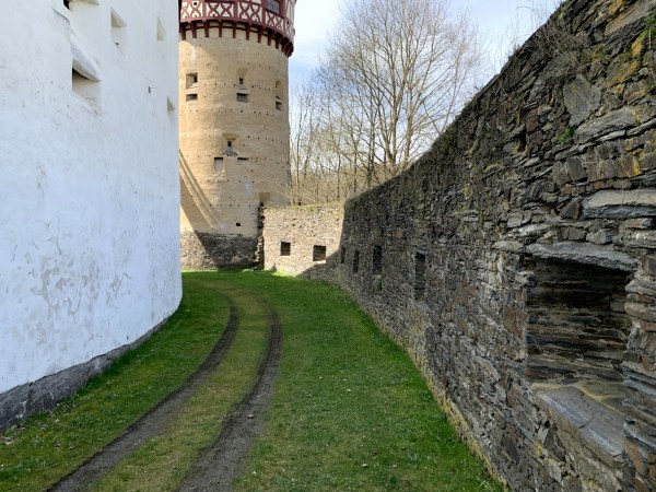 Nach links gebogener, grasbewachsener Weg mit Wagenspuren zwischen weißer Hausmauer und Steinmauer zum Turm.