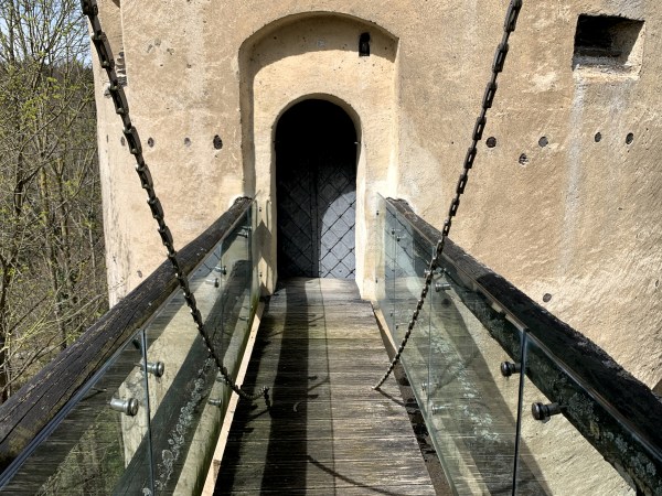 Holzweg auf einer Zugbrücke zu einer Tür in einem Turm. Die Brücke hat Holzgeländer die mit Glaswänden gesichert sind.