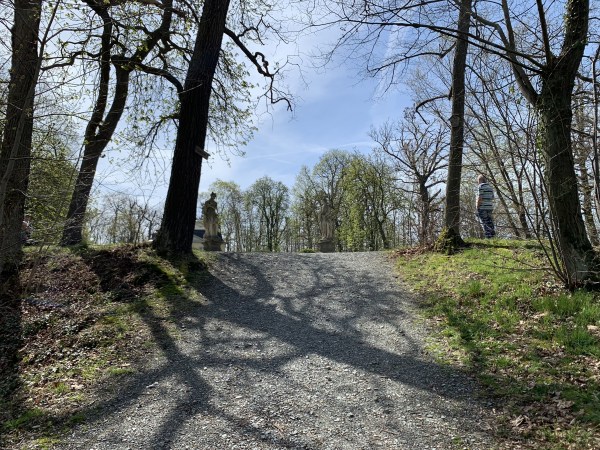 Weg zwischen Bäumen nach oben. Oben stehen Statuen und ein Mann