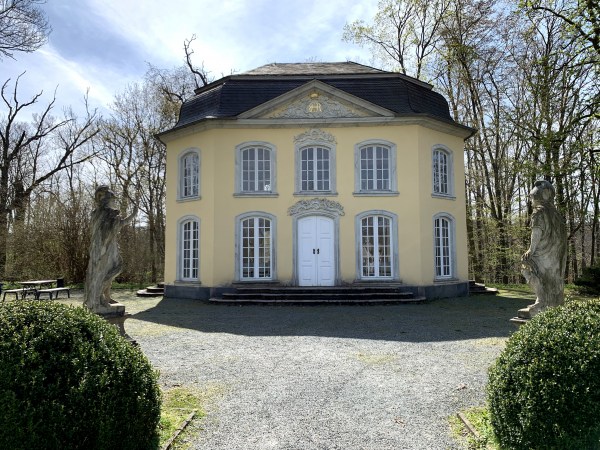 Zweistöckiges Gebäude mit beiger Wandfarbe zwischen zwei Statuen vor Bäumen. Das Häuschen hat große weiße Fenster. Der umgebene Park ist gepflegt, zu sehen an beschittenen Büschen und einem Schotterweg.