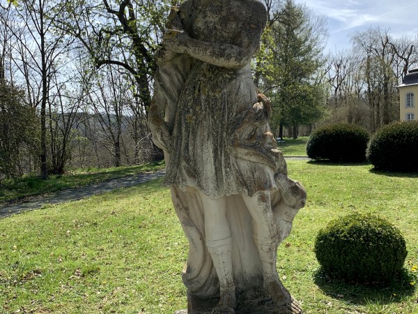 Weiße Statue einer Frau im Park vor gemähtem Rasen. Die Statue hat bereits grünbraune Altersflecken. Das Licht scheint von hinten.